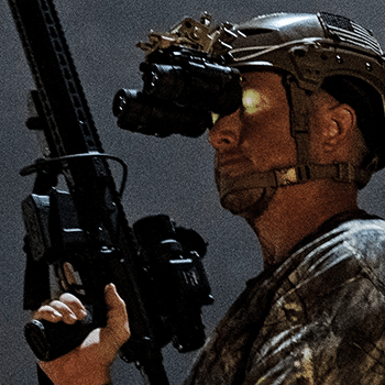 Man shown looking through a pair of helmet mounted thermal binoculars while holding an AR-15 style rifle with a Trijicon REAP-IR attached.